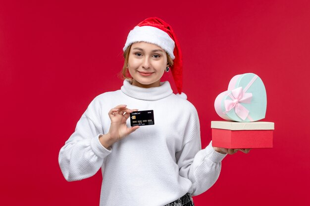 Vue de face jeune femme avec des cadeaux et une carte bancaire sur le fond rouge