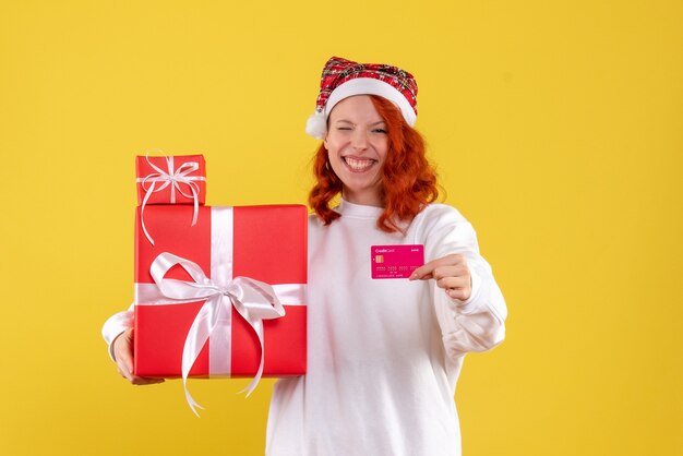 Vue de face de la jeune femme avec des cadeaux de Noël et carte bancaire sur mur jaune