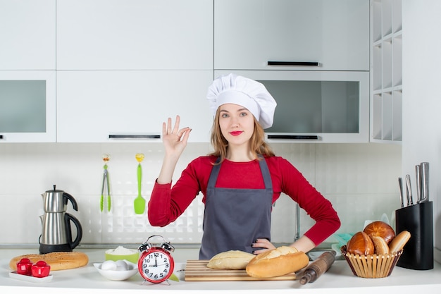 Vue de face jeune femme en chapeau de cuisinier et tablier debout derrière la table dans la cuisine