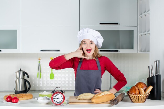 Vue de face jeune femme en chapeau de cuisinier et tablier faisant appelez-moi signe dans la cuisine