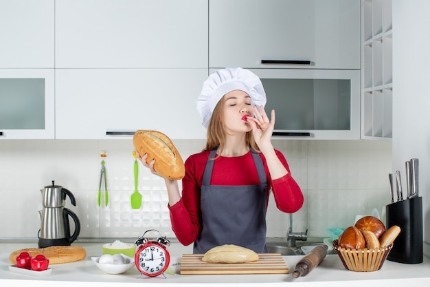 Vue de face jeune femme en chapeau de cuisinier et tablier tenant du pain faisant un baiser en chef dans la cuisine