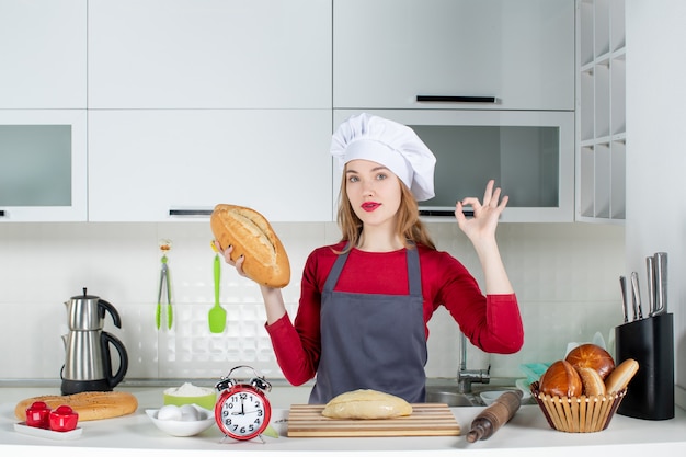 Vue de face jeune femme en chapeau de cuisinier et tablier tenant du pain faisant signe okey dans la cuisine