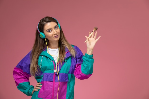 Vue de face jeune femme en chemise blanche et manteau coloré tenant un pinceau de maquillage sur le mur rose, modèle femme