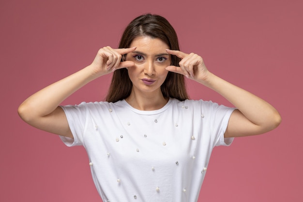 Vue de face jeune femme en chemise blanche montrant ses yeux avec les doigts sur le mur rose clair, modèle femme pose
