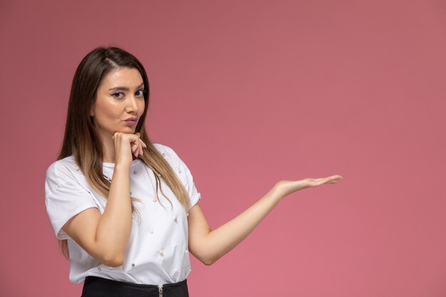 Vue de face jeune femme en chemise blanche posant avec la main levée sur le mur rose, modèle femme couleur posant femme