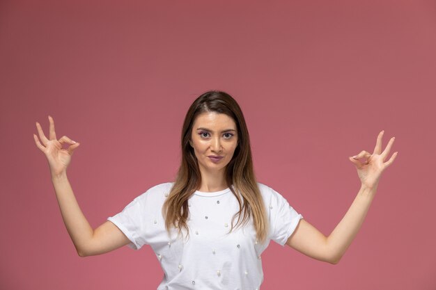 Vue de face jeune femme en chemise blanche posant en méditation pose sur mur rose, modèle de pose de femme de couleur