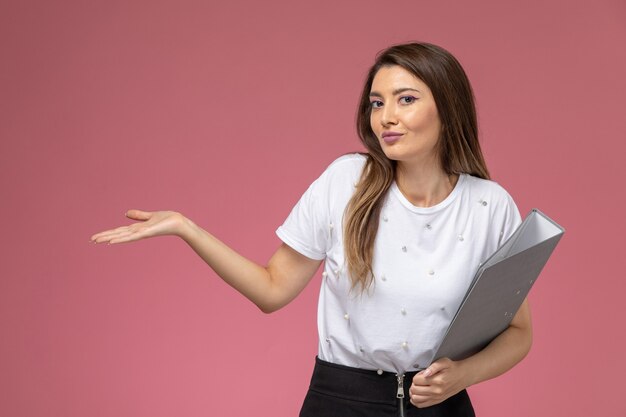 Vue de face jeune femme en chemise blanche tenant des fichiers gris sur un mur rose clair, femme modèle femme pose