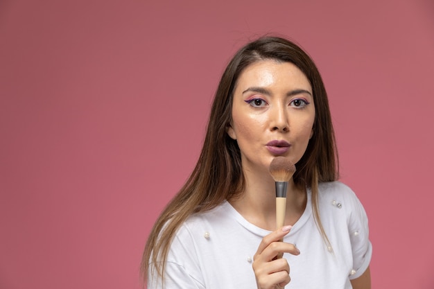 Vue de face jeune femme en chemise blanche tenant un pinceau de maquillage sur le mur rose, modèle de pose de femme de couleur