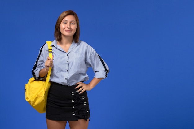 Vue de face de la jeune femme en chemise bleue portant un sac à dos jaune posant et souriant sur le mur bleu