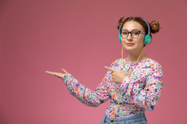 Vue de face jeune femme en chemise conçue de fleurs et blue-jeans, écouter de la musique avec un casque sur le bureau rose