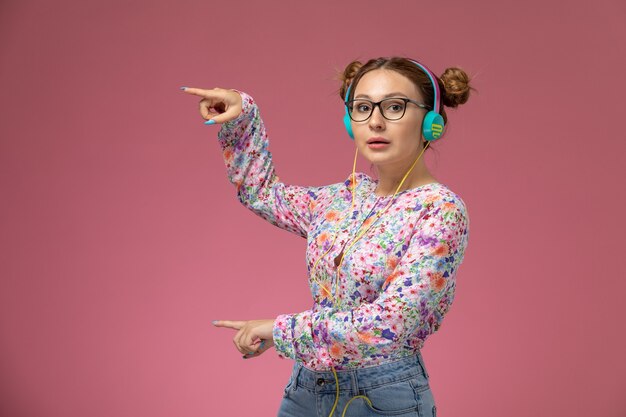 Vue de face jeune femme en chemise conçue de fleurs et blue-jeans, écouter de la musique avec des écouteurs sur fond rose