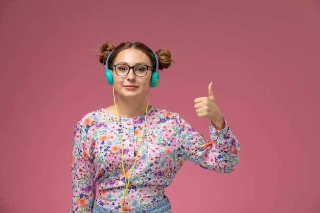 Vue de face jeune femme en chemise conçue de fleurs et blue-jeans, écouter de la musique avec le sourire sur le fond rose