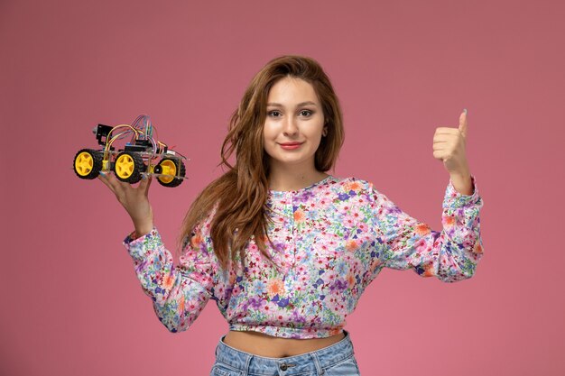 Photo gratuite vue de face jeune femme en chemise conçue de fleurs et blue-jeans holding toy car smiling sur fond rose