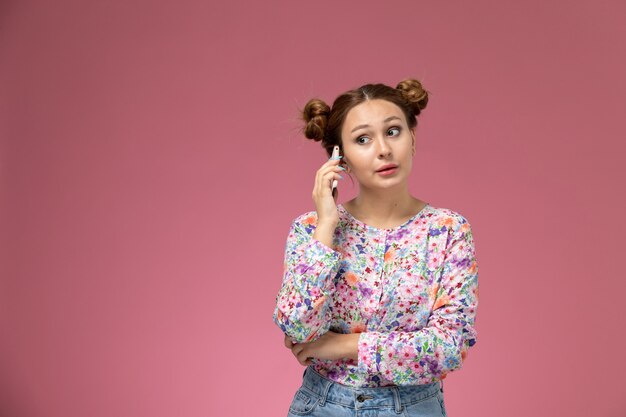 Vue de face jeune femme en chemise conçue de fleurs et blue-jeans parlant au téléphone sur le fond rose