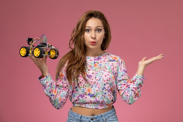 Vue de face jeune femme en chemise conçue de fleurs et blue-jeans pensant et tenant la voiture jouet sur le fond rose