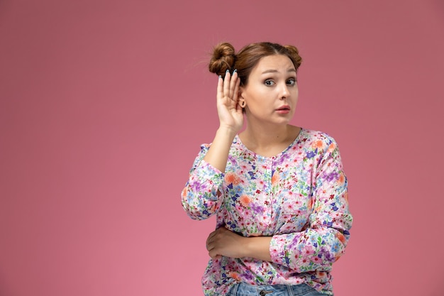 Vue De Face Jeune Femme En Chemise Conçue De Fleurs Et Jeans Bleu Essayant D'entendre Sur Fond Rose