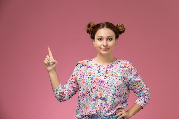 Vue de face jeune femme en chemise conçue de fleurs et jeans bleu posant avec le doigt sur le fond rose