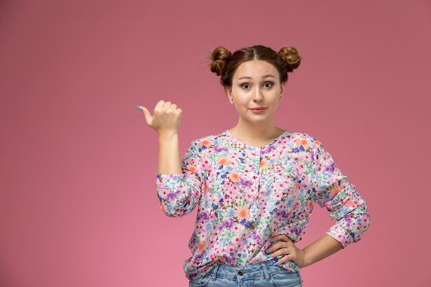 Vue de face jeune femme en chemise conçue de fleurs et jeans bleu posant avec la main levée sur le fond rose