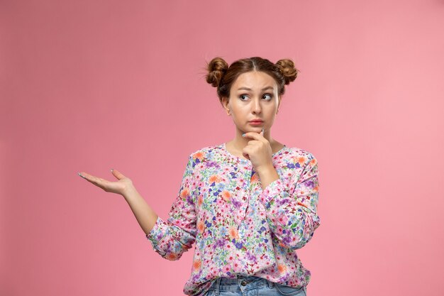 Vue de face jeune femme en chemise conçue de fleurs et jeans bleu posant avec la pensée du doigt sur le fond rose