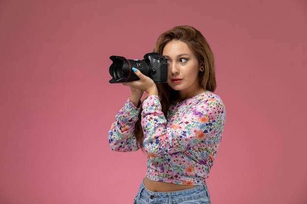 Photo gratuite vue de face jeune femme en chemise conçue de fleurs et jeans bleu prenant une photo avec appareil photo sur le fond rose