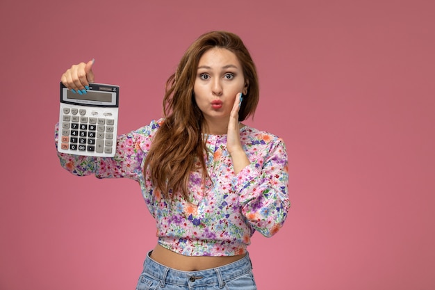 Vue de face jeune femme en chemise conçue de fleurs et jeans bleus tenant calculatrice sur bureau rose