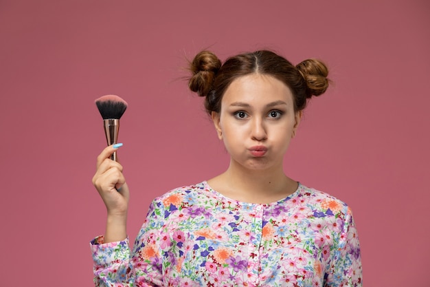 Vue de face jeune femme en chemise conçue de fleurs tenant un pinceau de maquillage sur le fond rose