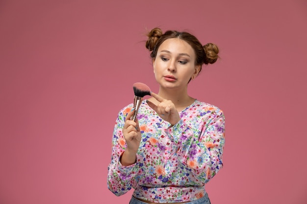 Vue de face jeune femme en chemise conçue de fleurs tenant un pinceau de maquillage sur le fond rose