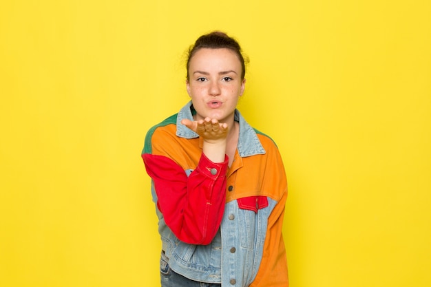 Photo gratuite une vue de face jeune femme en chemise jaune veste colorée et blue-jeans posant et envoyant des bisous aériens