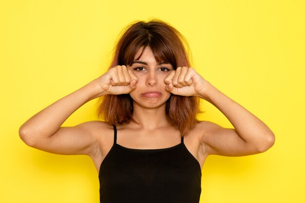 Vue de face de la jeune femme en chemise noire et faux jeans gris pleurant sur le mur jaune