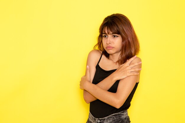 Vue de face de la jeune femme en chemise noire et jeans gris frissonnant sur mur jaune