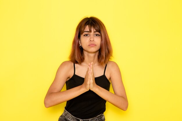 Vue de face de la jeune femme en chemise noire et jeans gris posant sur un mur jaune clair