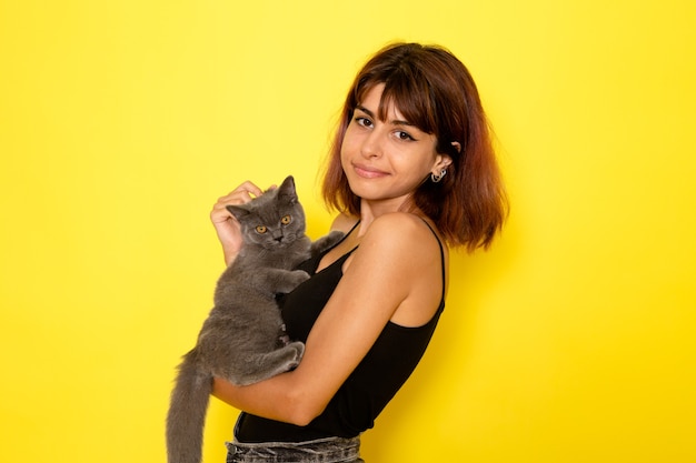 Vue de face de la jeune femme en chemise noire et jeans gris tenant chaton gris sur mur jaune