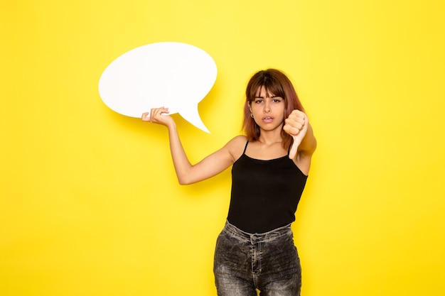 Photo gratuite vue de face de la jeune femme en chemise noire et jeans gris tenant énorme panneau blanc sur mur jaune