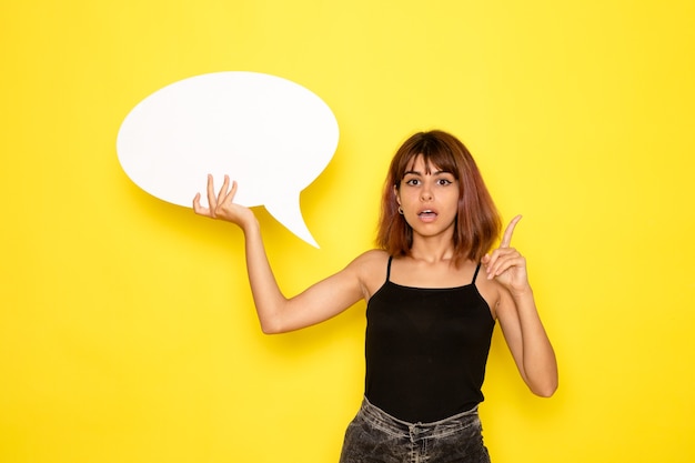 Photo gratuite vue de face de la jeune femme en chemise noire et jeans gris tenant grand panneau blanc sur mur jaune clair