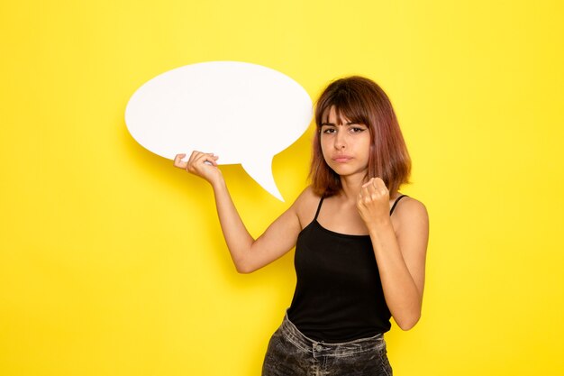 Vue de face de la jeune femme en chemise noire et jeans gris tenant grand panneau blanc sur mur jaune