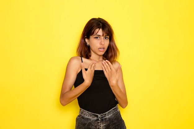 Vue de face de la jeune femme en chemise noire posant sur le mur jaune clair