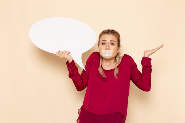 Vue de face jeune femme en chemise rouge avec bouche attachée tenant un énorme signe blanc sur l'espace crème femme tissu photo violence domestique