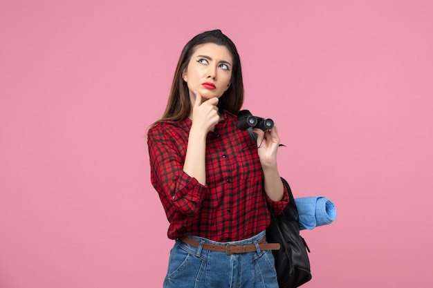 Vue de face jeune femme en chemise rouge avec des jumelles sur fond rose couleur femme étudiante