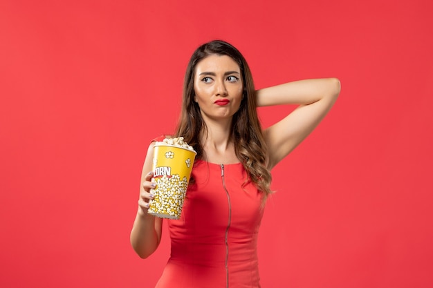 Vue de face jeune femme en chemise rouge tenant du pop-corn et penser sur la surface rouge