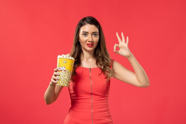 Vue de face jeune femme en chemise rouge tenant du pop-corn et souriant sur une surface rouge