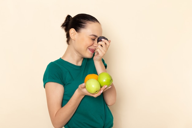 Vue de face jeune femme en chemise vert foncé et blue-jeans tenant différents fruits sur beige