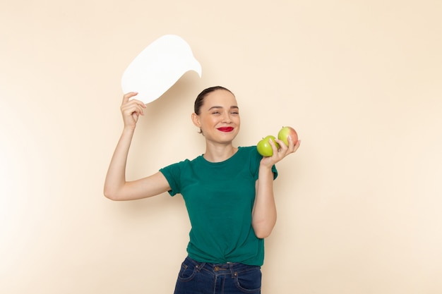 Vue de face jeune femme en chemise vert foncé et jean bleu tenant des pommes et panneau blanc sur beige