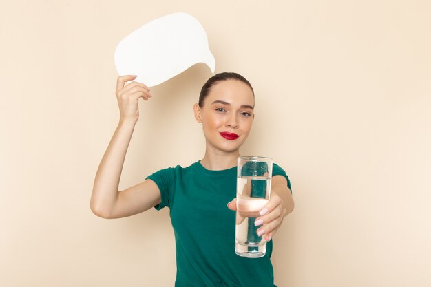 Vue de face jeune femme en chemise vert foncé et jean bleu tenant un verre d'eau et panneau blanc sur beige