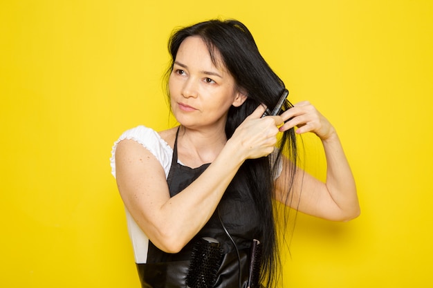 Une vue de face jeune femme coiffeuse en t-shirt blanc cape noire avec des brosses fixant ses cheveux vague de cheveux