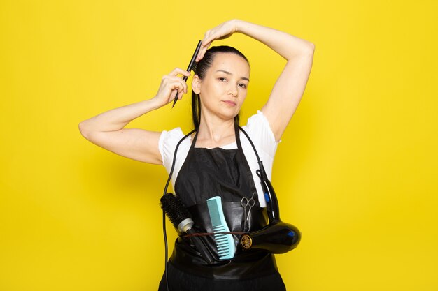 Une vue de face jeune femme coiffeuse en t-shirt blanc cape noire avec des brosses et un sèche-cheveux se brosser les cheveux posant