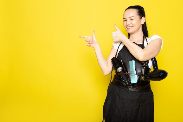 Une vue de face jeune femme coiffeuse en t-shirt blanc cape noire avec des brosses et un sèche-cheveux souriant posant accueillant