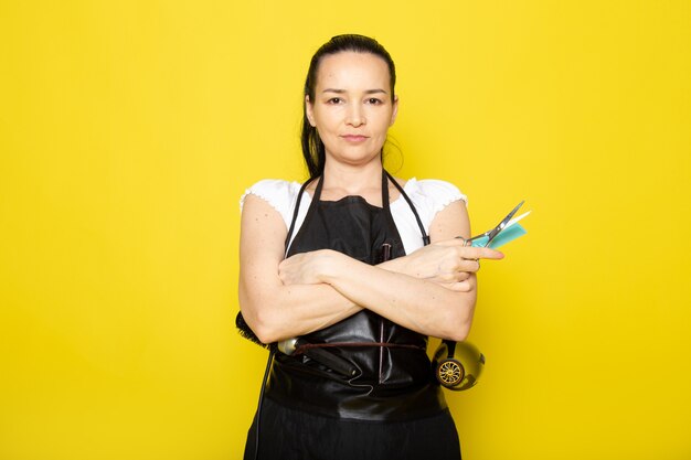 Une vue de face jeune femme coiffeuse en t-shirt blanc cape noire avec des brosses et un sèche-cheveux tenant des ciseaux posant