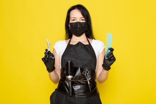 Une vue de face jeune femme coiffeuse en t-shirt blanc cape noire tenant une brosse à cheveux bleue et des ciseaux en masque noir stérile gants noirs