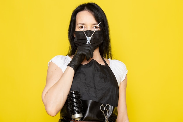 Une vue de face jeune femme coiffeuse en t-shirt blanc cape noire tenant des ciseaux en masque noir stérile gants noirs