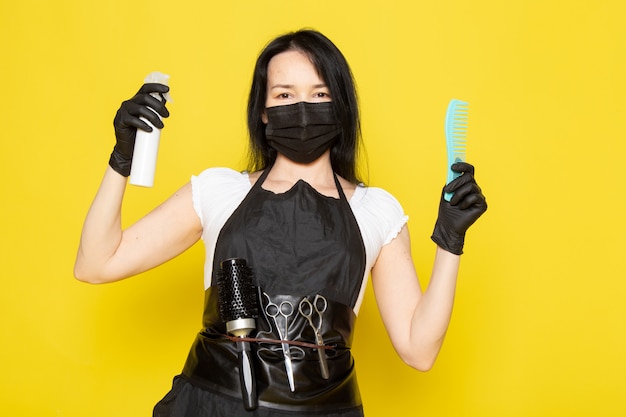 Une vue de face jeune femme coiffeuse en t-shirt blanc cape noire tenant un spray et une brosse à cheveux en masque noir stérile gants noirs